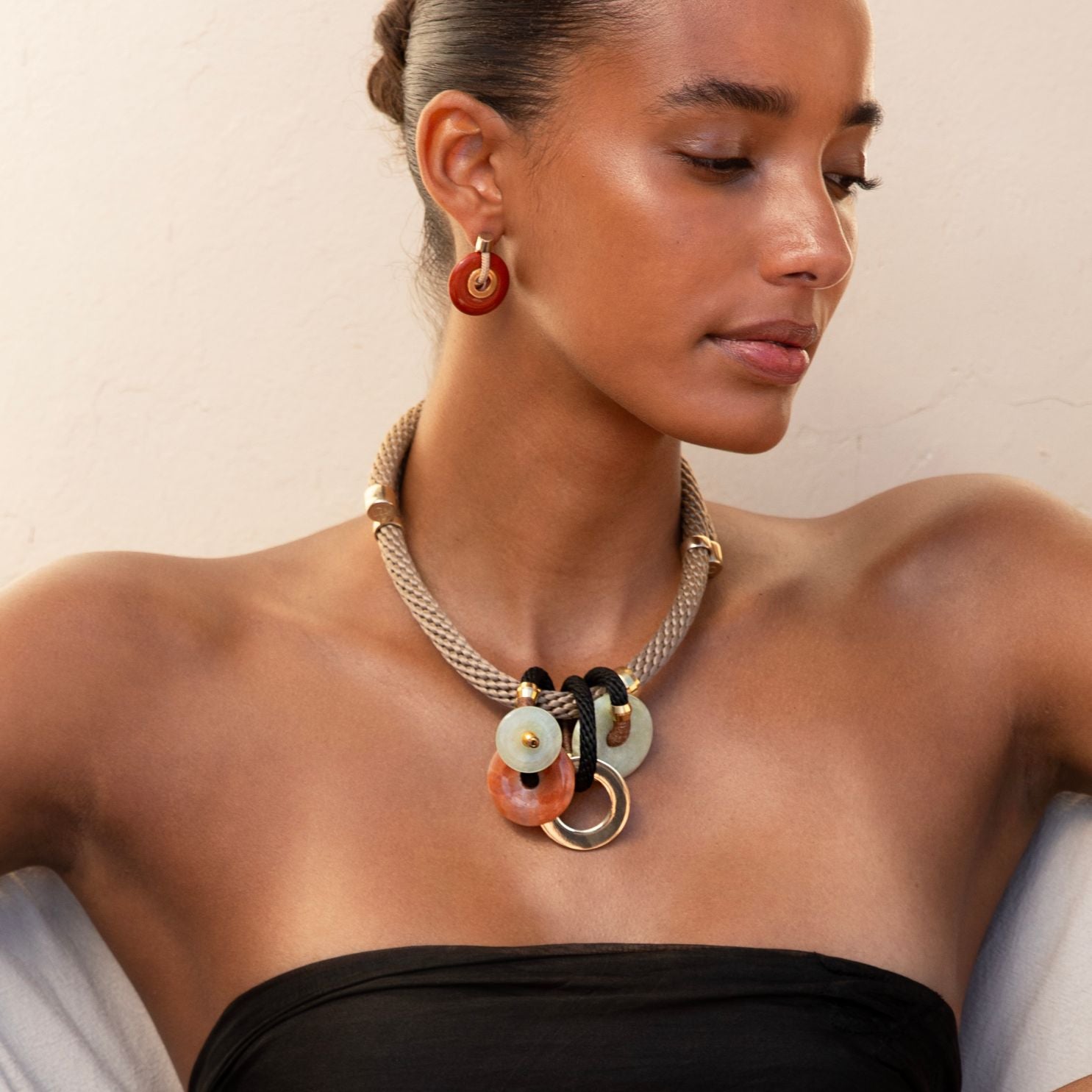 A braided beige rope necklace with gold-toned hardware and three dangling charms: two pale green circular discs, one terracotta orange circular disc, and a small gold ring, shown on a light gray background.